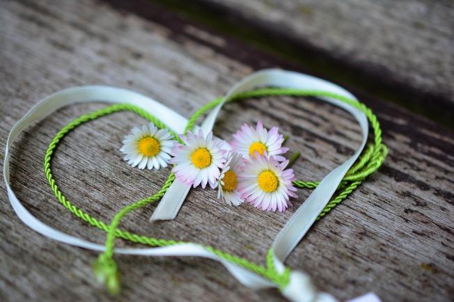 A heart made of green and white ribbon, decorated with small daisies, is placed on wood.
